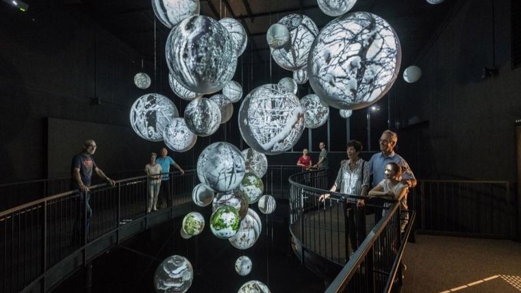 Eine moderne Ausstellung mit schwebenden, beleuchteten Kugeln, die Naturmotive zeigen. Besucher beobachten die Installation in einem dunklen Raum.