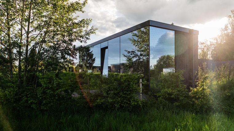 Ein modernes, gläsernes Gebäude umgeben von grünen Bäumen und Sträuchern. Die Architektur harmoniert mit der natürlichen Umgebung.