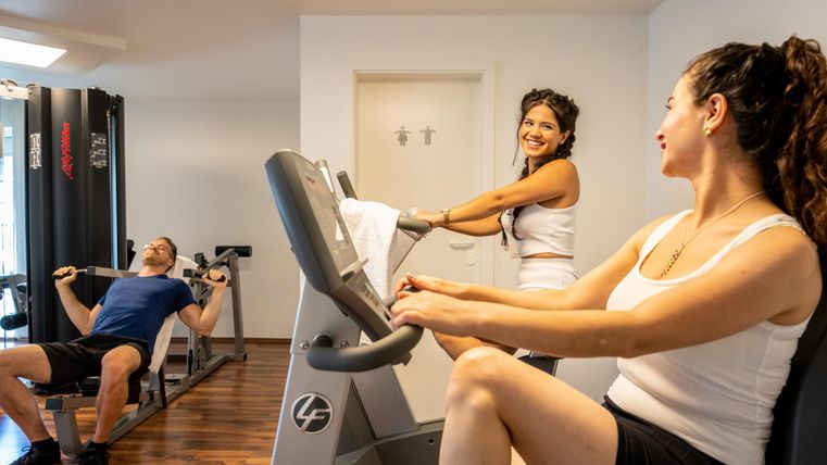 Ein modernes Fitnessstudio mit drei Personen beim Training. Zwei Frauen benutzen Cardio-Geräte, während ein Mann an einer Gewichtmaschine trainiert.