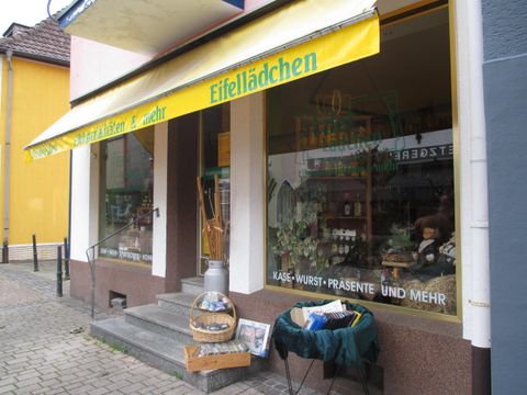 Ein kleines Geschäft mit einem gelben Schild "Eifellädchen". Die Fenster zeigen verschiedene Produkte und Accessoires.