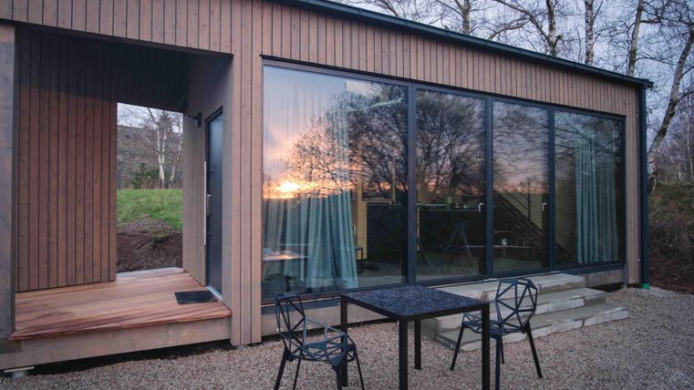 Ein modernes Holzhaus mit großen Fensterfronten, die den Sonnenuntergang reflektieren. Vor dem Haus steht ein Tisch mit Stühlen auf einem kieselgesäumten Platz.