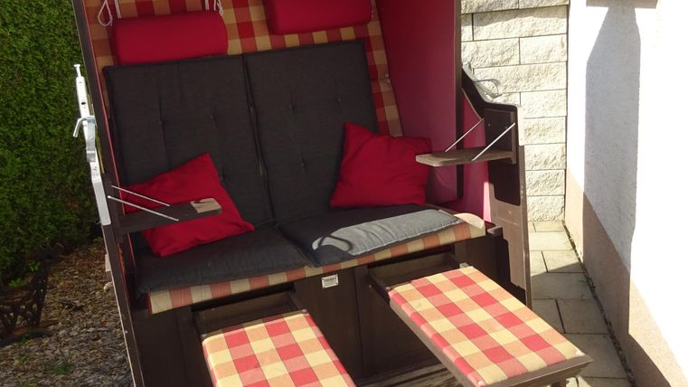 Ein gemütlicher Strandkorb mit roten Kissen steht auf einem gepflasterten Weg. Daneben befinden sich zwei kleine Tische mit karierter Tischdecke.