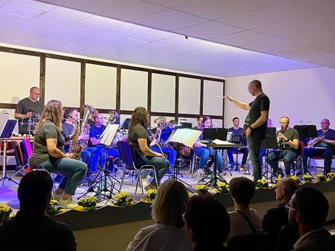 Ein Orchester spielt auf einer Bühne, während der Dirigent die Musiker anleitet. Verschiedene Instrumente sind zu sehen, und das Publikum sitzt im Vordergrund.