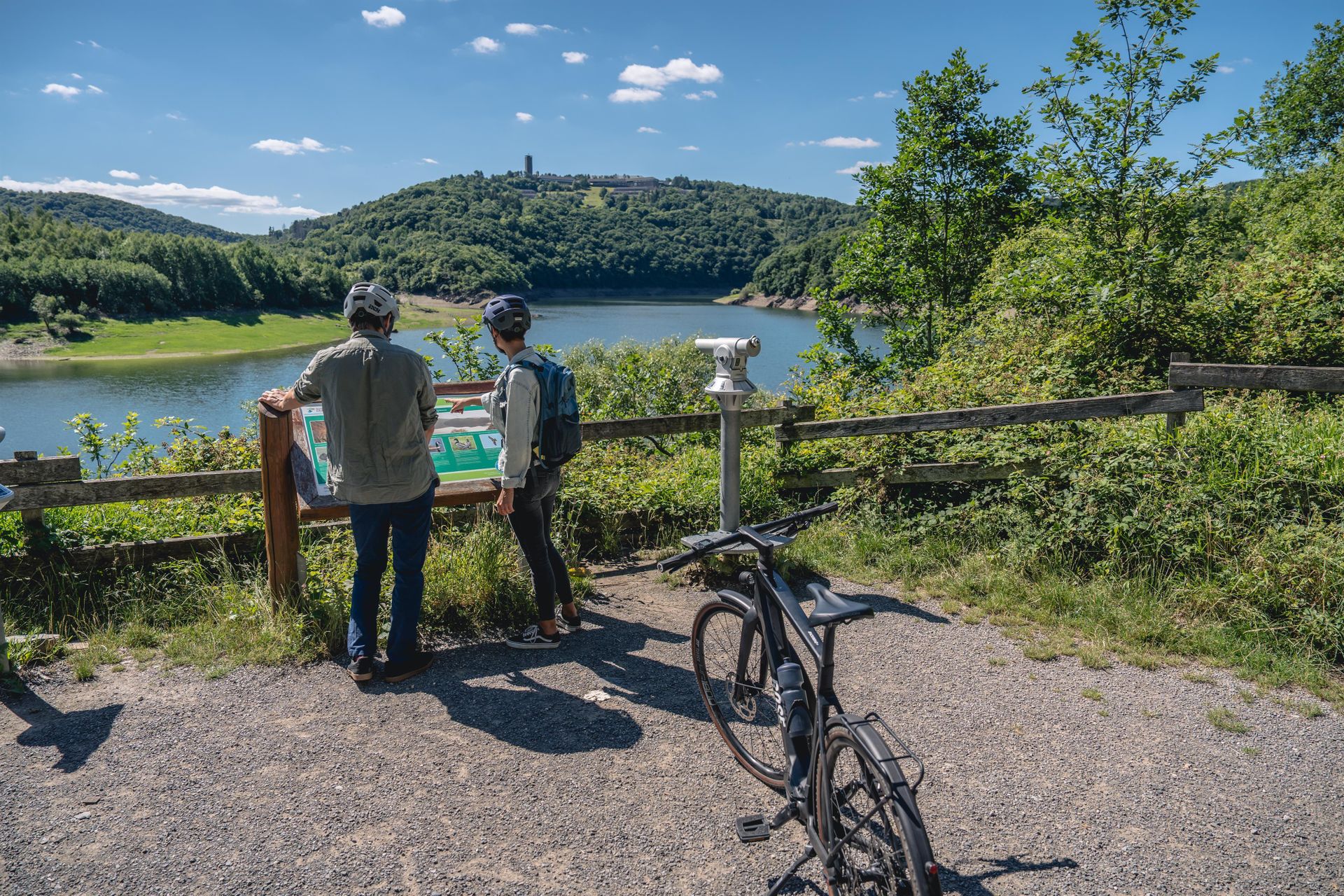 Zwei Personen stehen an einem Aussichtspunkt und betrachten eine Landschaft mit einem See und Hügeln. Ein Fahrrad steht neben ihnen auf dem Weg, umgeben von grüner Vegetation.