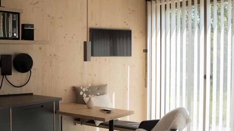 Ein modernes Zimmer mit Holzverkleidung, einem Tisch und einem Stuhl. An der Wand hängt ein Fernseher, und große Fenster mit Jalousien lassen viel Licht herein.