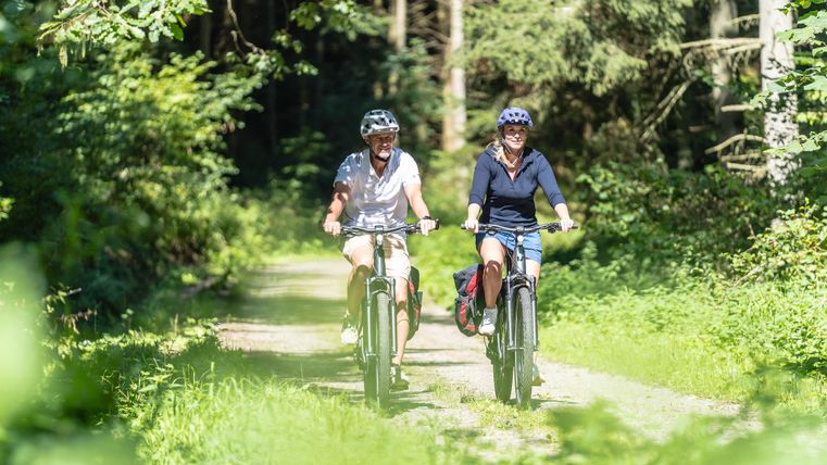 Zwei Personen fahren mit ihren E-Bikes auf einem Waldweg. Um sie herum ist üppige grüne Natur.