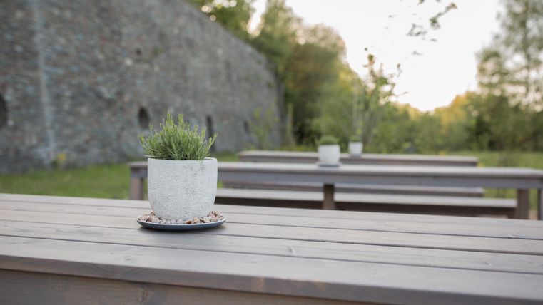 Ein gemütlicher Outdoor-Bereich mit Holztischen und einer Betonblume. Im Hintergrund ist eine alte Mauer und grüne Bäume zu sehen.