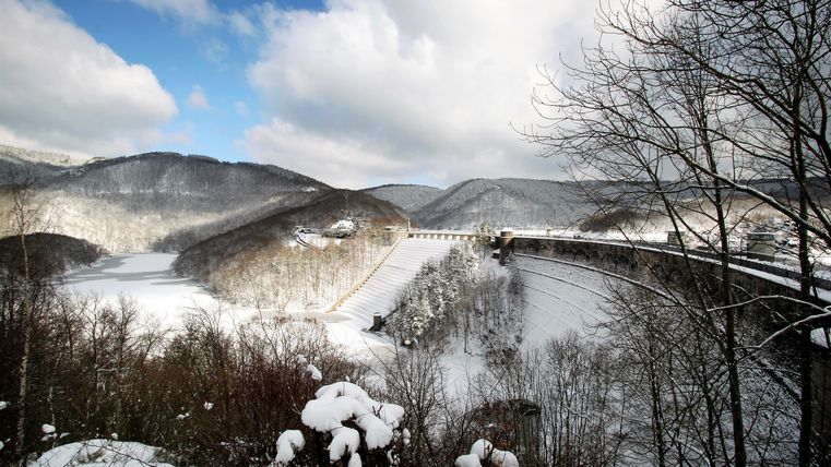 Eine winterliche Landschaft mit schneebedeckten Hügeln und einer Brücke. Der Himmel ist klar mit einigen Wolken.