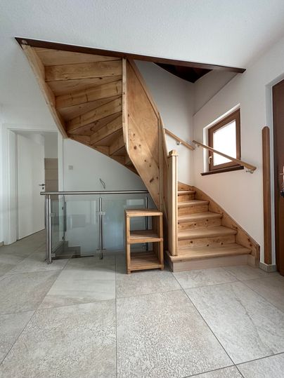 Eine elegante Treppe aus Holz, die in einen hellen Raum führt. Der Boden ist mit grauen Fliesen gestaltet und es gibt große Fenster, die für viel Licht sorgen.