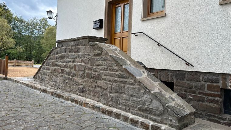 Ein Steinhang führt zur Eingangstür eines Hauses. Die Umgebung ist grün und es gibt einen gepflasterten Weg.