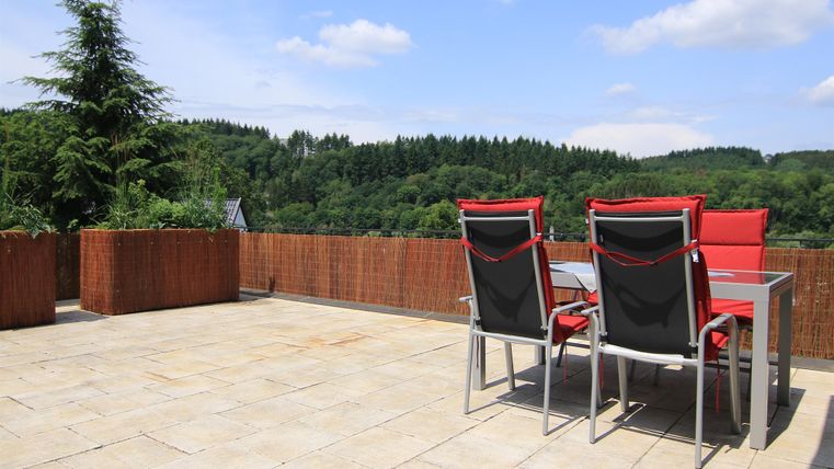 Ein geräumiger Terrassenbereich mit einem Tisch und Stühlen. Im Hintergrund sieht man eine grüne Waldlandschaft unter einem klaren blauen Himmel.