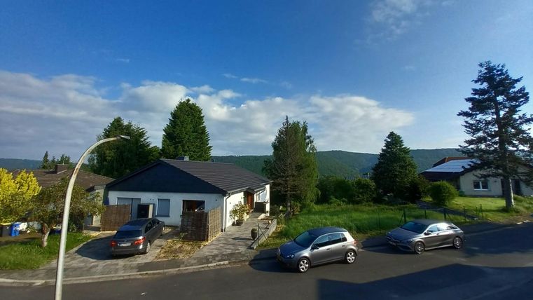 Ein ruhiges Wohngebiet mit mehreren Autos auf der Straße. Im Hintergrund sind grüne Hügel und ein blauer Himmel zu sehen.
