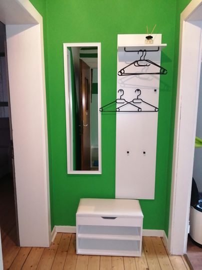 Ein Eingangsbereich mit grüner Wandfarbe, einem Spiegel und einer Garderobe. Es gibt auch eine weiße Sitzbank mit Stauraum.