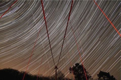 Ein beeindruckendes Sternenbild mit langem Belichtungszeitraum zeigt die Bewegungen der Sterne am Nachthimmel. Rote Linien von einer Brücke kreuzen das Bild und verleihen ihm zusätzliches Interesse.