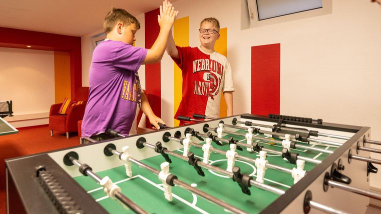 Zwei Jungen hoch fünf, nachdem sie miteinander am Tischfußball gespielt haben. Der Raum ist hell und modern eingerichtet.