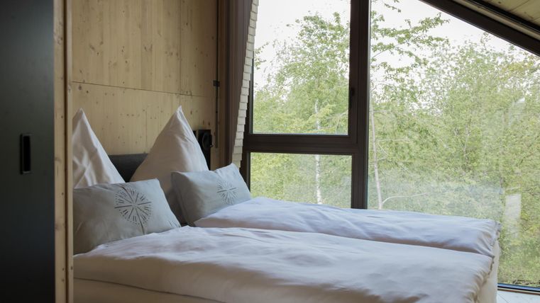 Ein modernes Schlafzimmer mit zwei Betten und großen Fenstern. Der Ausblick zeigt grüne Bäume und eine ruhige Umgebung.