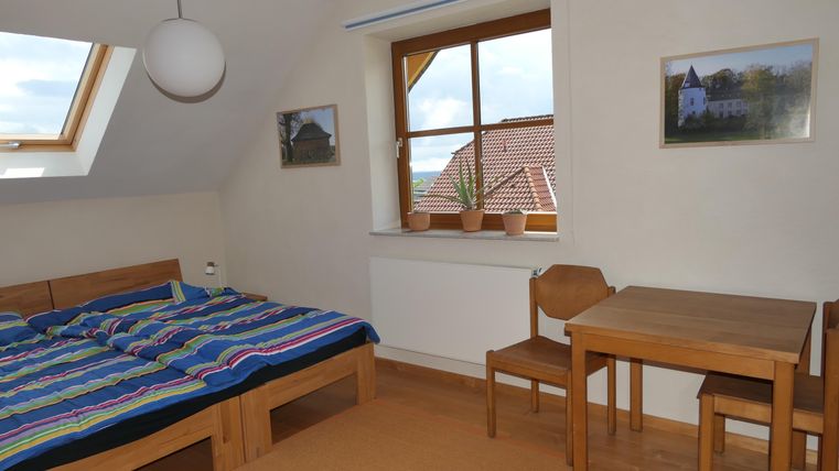 Ein gemütliches Zimmer mit einem Doppelbett und bunten Bettwäsche. Am Fenster gibt es Pflanzen, und ein Tisch mit Stühlen ist auf der gegenüberliegenden Seite.