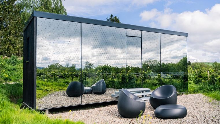Ein modernes, spiegelndes Gebäude steht in einer grünen Landschaft. Vor dem Gebäude befinden sich bequeme, schwarze Sitzmöbel.