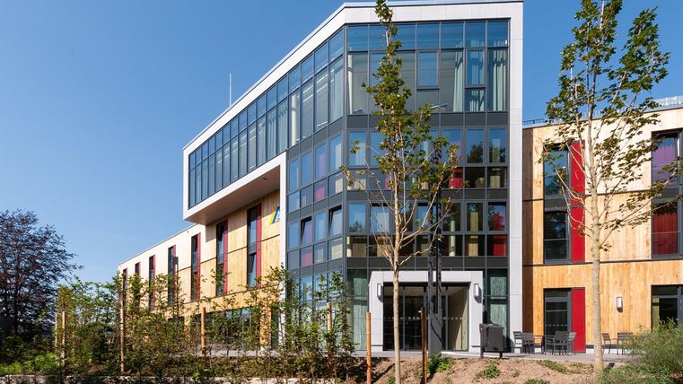 Ein modernes Gebäude mit großen Fenstern und Holzverkleidung. Umgeben von Bäumen und grüner Landschaft.