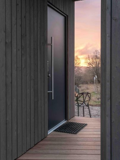 Ein moderner Eingang mit einer schwarzen Tür und einer Holzveranda. Im Hintergrund ist ein schöner Sonnenuntergang und einige Bäume sichtbar.