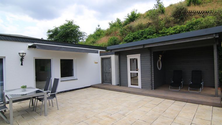 Ein modernes Haus mit einer Terrasse aus Steinplatten. Im Hintergrund sind grüne Hügel und ein überdachter Bereich mit Liegen sichtbar.