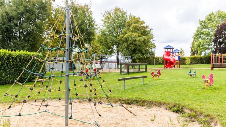 Ein Spielplatz mit einem Klettergerüst und einer Rutsche. Umgeben von grünen Bäumen und Wiesen.