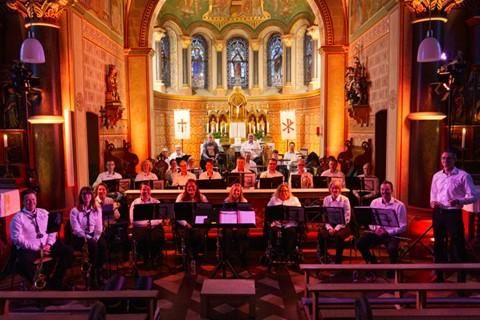 Eine Gruppe von Musikern sitzt in einer Kirche. Der Innenraum ist festlich beleuchtet und zeigt eine schöne Architektur.