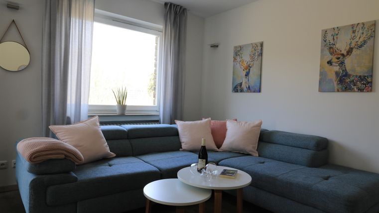 Ein modernes Wohnzimmer mit einem blauen Sofa und Kissen in sanften Farben. An den Wänden hängen zwei Gemälde, und ein kleiner Tisch steht in der Mitte.