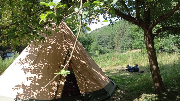 Ein Zelt steht in einer grünen Wiese, umgeben von Bäumen. Im Hintergrund sind zwei Personen zu sehen, die sich entspannen.