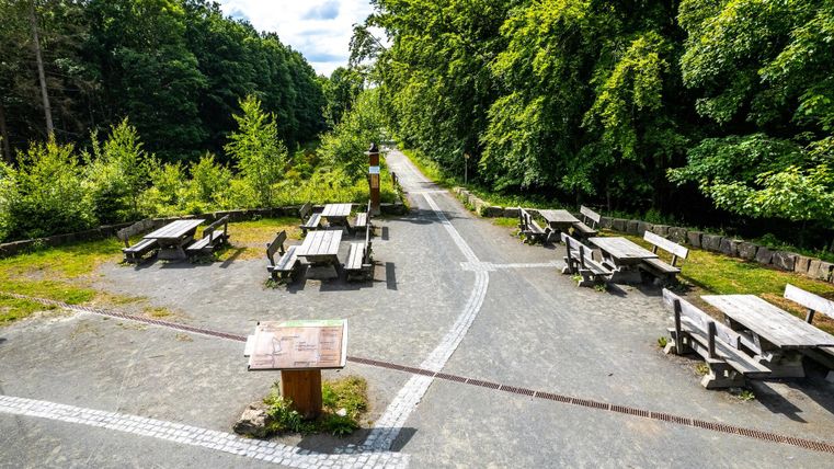 <p>Ein Rastplatz im Wald auf dem Tische und Bänke stehen.Vorne im Bild ist ein kleines Holzpult mit einer Informationstafel. Der Weg führt durch die Sitzmöglichkeiten in den Wald. Neben dem Weg steht Säule mit einem hölzernen Hut oben drauf. Das ist der Treffpunkt für Wanderungen.  </p>