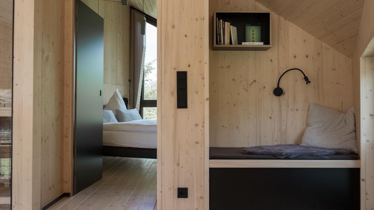 Ein modernes Zimmer mit Holzverkleidung und großen Fenstern. Auf der linken Seite ist ein Bett und auf der rechten Seite eine gemütliche Leseecke.