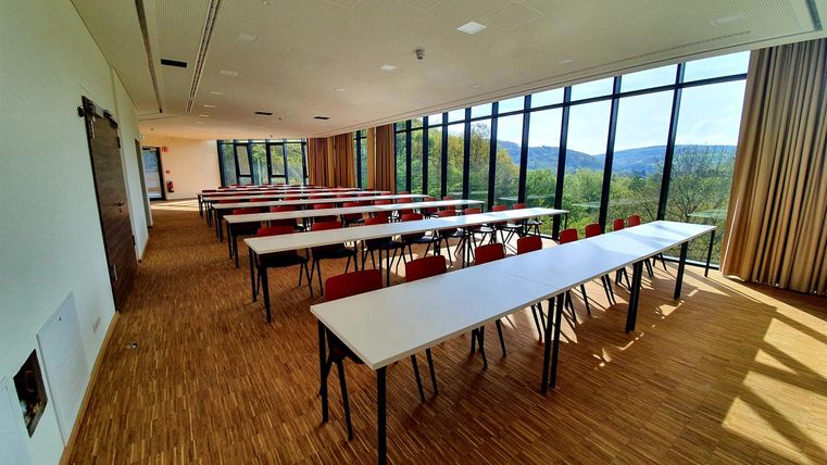 Ein moderner Seminarraum mit großen Fenstern und Blick auf die Landschaft. Es gibt mehrere Tische und Stühle, die für eine Gruppe bereitstehen.