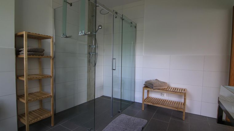 Ein modernes Badezimmer mit einer Glasdusche und minimalistischem Design. Auf der Seite steht ein Holzregal mit Handtüchern und eine Bank.