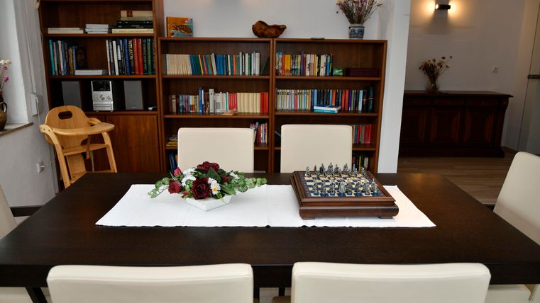 Ein eleganter Esstisch mit vier weißen Stühlen und einem Schachspiel. Im Hintergrund befindet sich ein Bücherregal mit einer Vielzahl von Büchern.