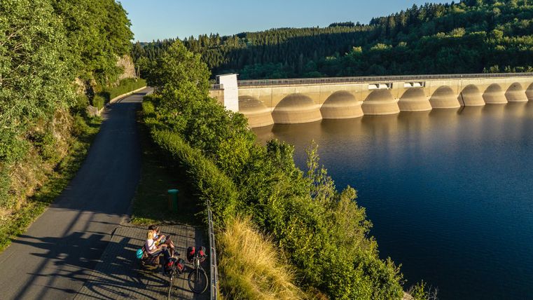 Ein malerisches Schloss umgeben von Grün und Bäumen. Radfahrer fahren auf einem Weg im Vordergrund.