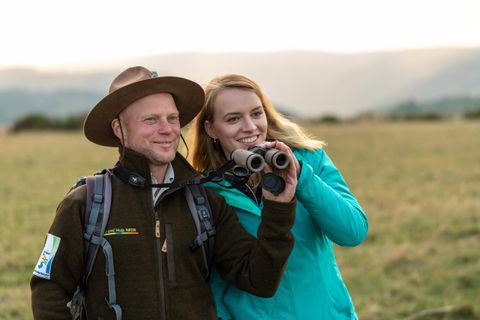 Ein Mann und eine Frau stehen in einem Feld. Sie halten ein Fernglas und lächeln, während sie die Landschaft beobachten.