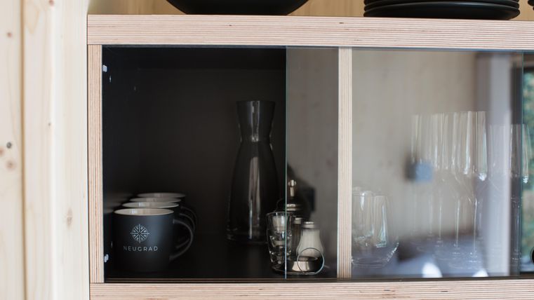 Ein moderner Küchenschrank mit Glastüren. Darin befinden sich Tassen, ein Krug und Gläser auf einem Holzregal.