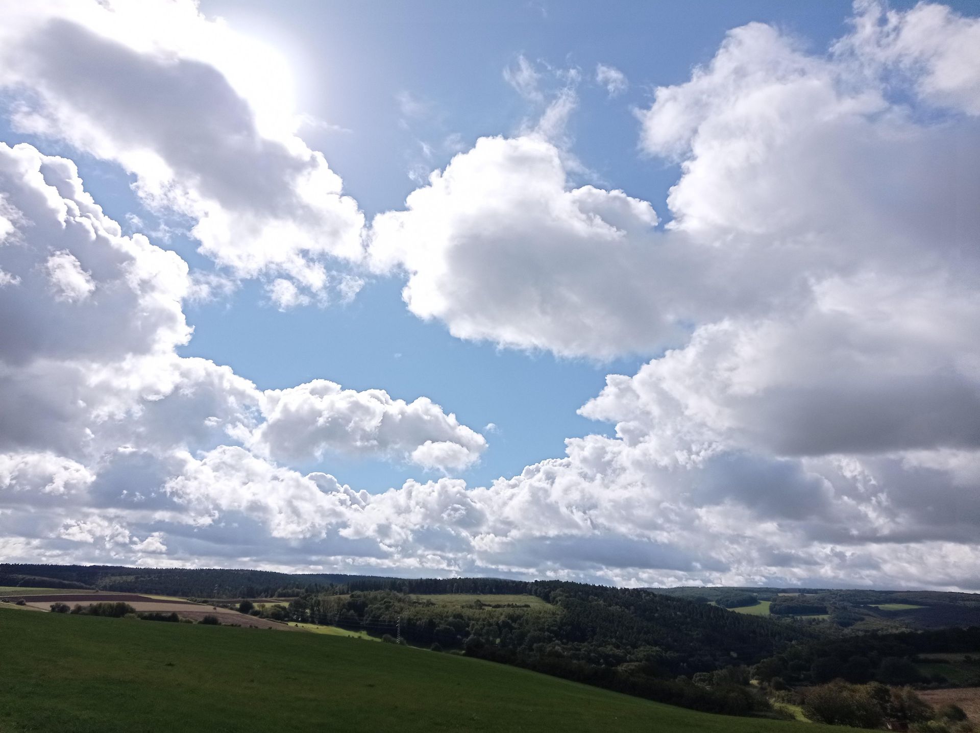 Ein klarer Himmel mit vielen Wolken und strahlendem Licht. Im Hintergrund sind sanfte Hügel und grüne Wiesen zu sehen.