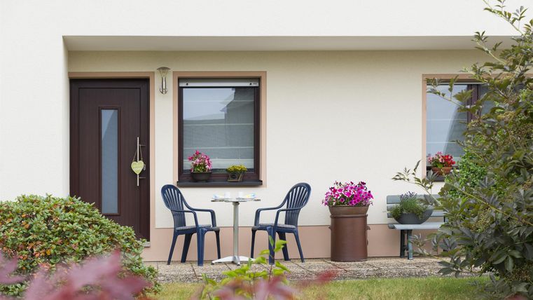 Eine gemütliche Terrasse mit zwei Stühlen und einem kleinen Tisch. Blumen sind in Töpfen vor den Fenstern und im Garten sichtbar.