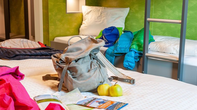 Ein gemütliches Zimmer mit zwei Betten und farbigen Jacken. Auf dem Bett liegt ein Rucksack und zwei Äpfel.