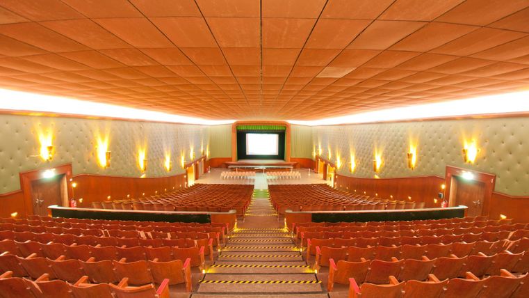 Ein lichtdurchflutetes Kino mit vielen roten Sitzen und einem großen Bildschirm. Die Wände sind in sanften Farbtönen gehalten, und die Treppen führen zum Zentrum des Raumes.