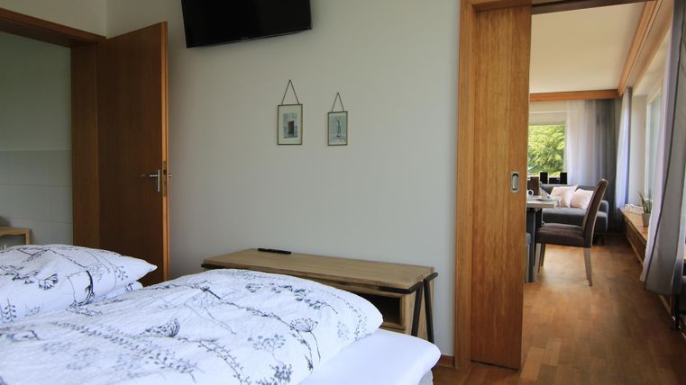 Ein gemütliches Schlafzimmer mit zwei Betten und einem Fernseher. Im Hintergrund ist ein heller Wohnbereich mit einer Essgruppe sichtbar.