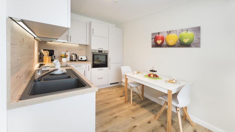 Eine moderne Küche mit einem Esstisch und hellen Möbeln. An der Wand hängen Bilder von buntem Obst.