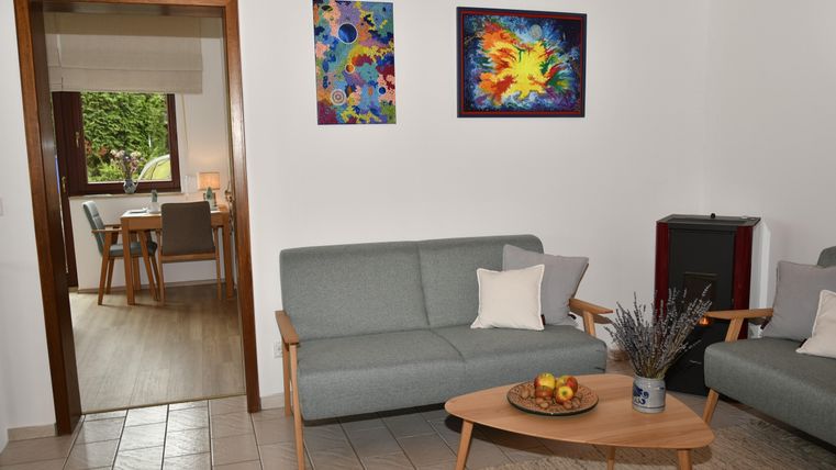 Ein helles Wohnzimmer mit zwei grauen Sofas und einem Holztisch. An der Wand hängen bunte Bilder, und im Hintergrund ist ein Essbereich sichtbar.