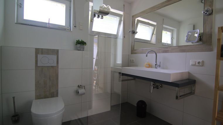 Ein modernes Badezimmer mit weißen Fliesen und einem großen Spiegel. Es gibt ein Waschbecken, eine Toilette und viel natürliches Licht durch die Fenster.