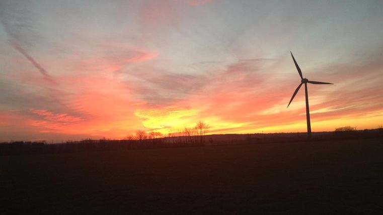 Eine Windkraftanlage steht im Vordergrund vor einem bunten Sonnenuntergang. Der Himmel ist in warmen Farben von Orange und Rosa gefärbt.