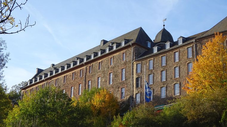 Ein historisches Gebäude mit mehreren Etagen, umgeben von grünen Bäumen und buntem Herbstlaub. Der Himmel ist klar und blau, was eine angenehme Atmosphäre schafft.