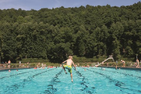 Een zwembad met een jongen die in het water springt. Op de achtergrond zijn andere zwemmers en groene bomen te zien.