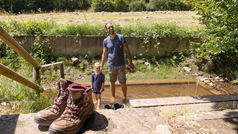 Ein Vater steht mit seinem Kind an einem kleinen Gewässer. Im Vordergrund liegen große Wanderschuhe.
