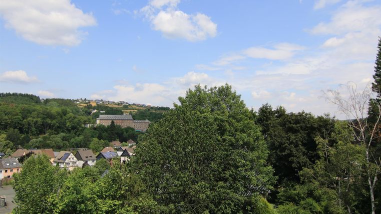 Eine grüne Landschaft mit Bäumen und einem Blick auf kleine Häuser. Der Himmel ist blau mit einigen Wolken.